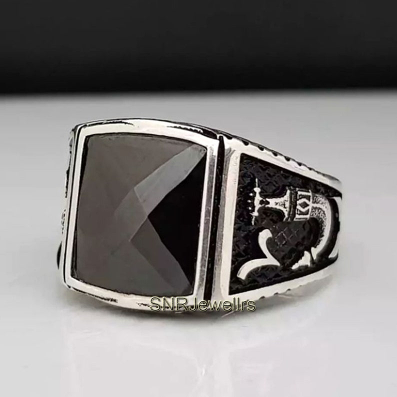 May include: A silver ring with a large, square, black gemstone. The ring has a detailed design on the sides, and the text "SNRJewellrs" is visible on the band.