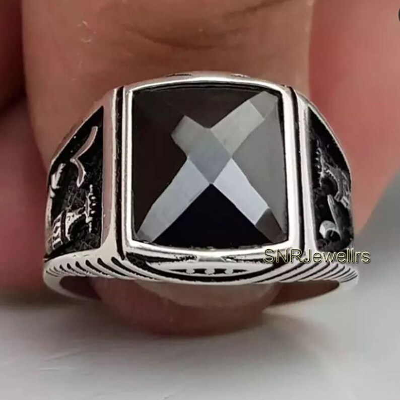 May include: A silver ring with a large, square, black gemstone. The ring has intricate side detailing and a ridged band. The text "SNRJewellrs" is visible on the ring.