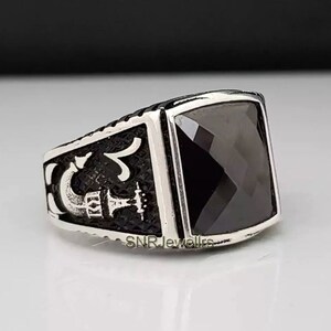 May include: A silver-toned ring with a large, square, black gemstone. The ring has a detailed design on the sides, with the text "SNRJewellrs" visible. The ring is likely made of metal and features a polished finish.