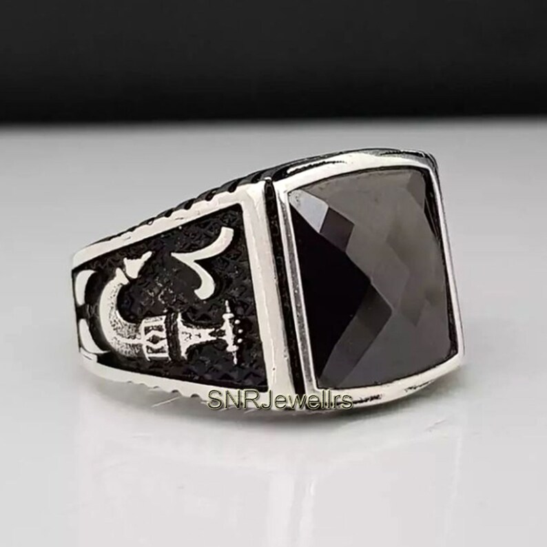 May include: A silver-toned ring with a large, square, black gemstone. The ring has a detailed design on the sides, with the text "SNRJewellrs" visible. The ring is likely made of metal and features a polished finish.