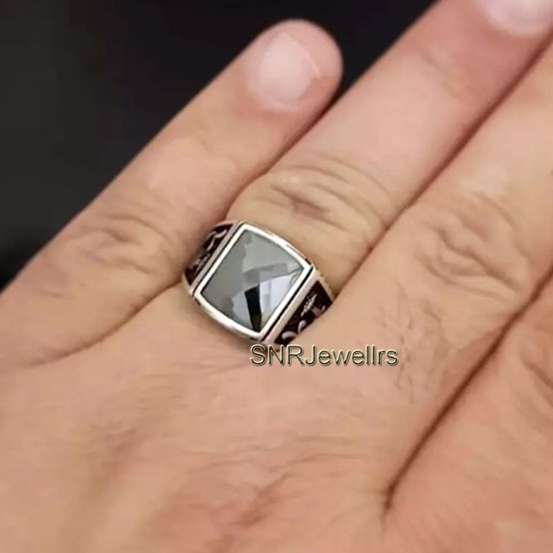 May include: A silver ring with a large, square, black gemstone. The ring has decorative black accents on the sides. The text "SNRJewellrs" is visible on the ring.