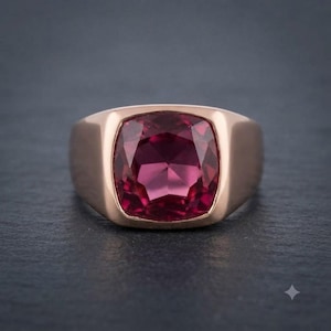 Könnte beinhalten: Ein Ring aus Roségold mit einem großen, kissenförmigen, tief magentafarbenen Edelstein in der Mitte. Die Ringschiene ist glatt und massiv, mit einer polierten Oberfläche, die den facettierten Stein ergänzt. Das Design ist schlicht und betont den Kontrast zwischen dem Metall und dem leuchtenden, dunkelrosa Edelstein.