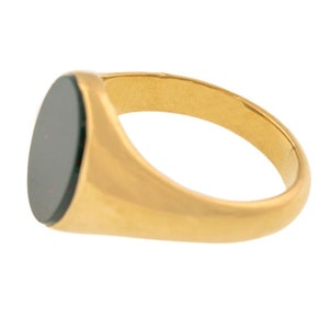 K&ouml;nnte beinhalten: Ein goldener Siegelring mit einem dunklen, ovalen Stein. Der Ring hat ein glattes, poliertes Band und eine flache, ovale Oberfl&auml;che. Der Stein hat eine dunkle Farbe, m&ouml;glicherweise schwarz oder tiefgr&uuml;n. Der Ring wird vor wei&szlig;em Hintergrund gezeigt.