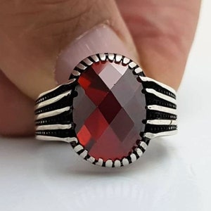 May include: A silver ring with a large, faceted, oval-shaped, deep red gemstone. The ring has a detailed design with horizontal lines on the band and a textured border around the gemstone. The ring is held against a white background.