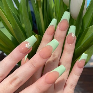 May include: Close-up of hands with long, light green and nude nails. The nails have a light green tip with a white flower accent.