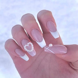 May include: Close-up of a hand with long, light pink nails with white tips. Some nails have small pearl accents and one nail has a pearl heart design.