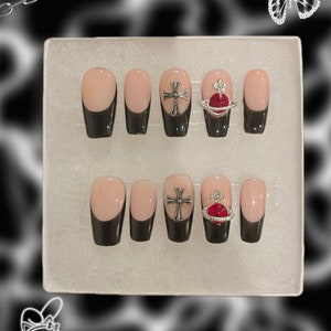 May include: A set of press-on nails with a black French tip design and a nude base. Some nails feature silver cross and red heart-shaped embellishments. The nails are displayed in a white box against a black and white background.