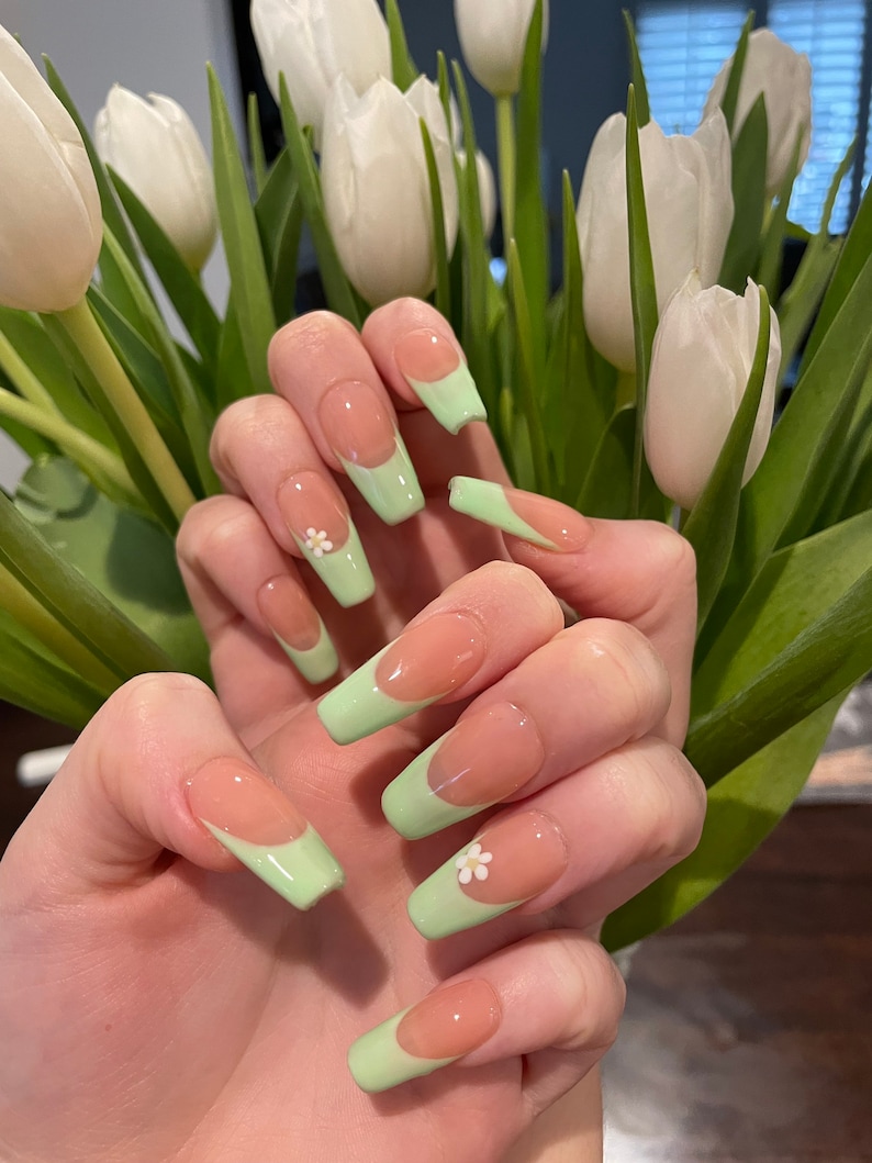 May include: Close-up of a hand with long, light pink, almond-shaped nails with a light green tip and a small white flower accent on some nails.