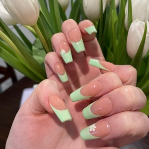 May include: Long, light green and nude colored fake nails with a white flower accent on one nail.