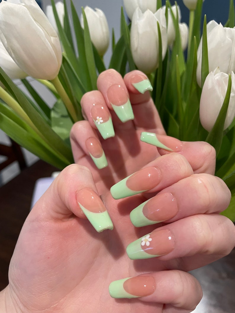 May include: Long, light green and nude colored fake nails with a white flower accent on one nail.