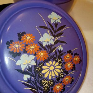 May include: A vintage purple cosmetic compact with a floral design. The lid features orange, yellow, and white flowers with black and brown stems and leaves. The compact is round and has a matching base.