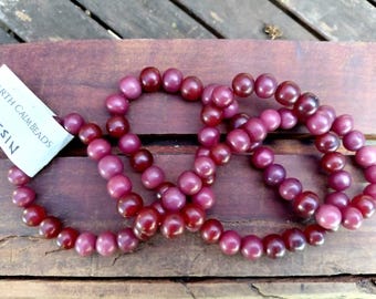 Puede incluir: Tres hilos de cuentas de resina granate se disponen sobre una superficie de madera. Las cuentas son redondas y brillantes. Se ve una etiqueta blanca con el texto "EARTH CALM BEADS RESIN".