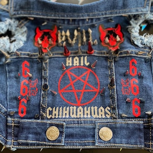 Puede incluir: Una chaqueta vaquera azul con un pentagrama rojo y las palabras "Hail Chihuahuas" en purpurina plateada. La chaqueta está decorada con parches de diablo rojos, tachuelas plateadas y el número 666.