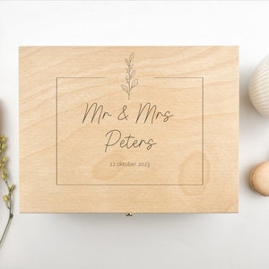 May include: A wooden box with a light brown finish. The box has a hinged lid and a small metal latch. The top of the box is engraved with a simple floral design, the text "Mr & Mrs Peters", and the date "12 oktober 2023".