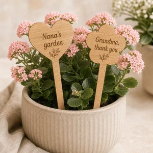 Op de afbeelding: Een lichtbeige geribbelde keramische pot met een groene plant met kleine roze bloemen. Twee houten plantenstekers zitten in de aarde: een hartvormige met de tekst "Nana's garden" en een bloemvormige met "Grandma, thank you".