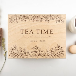 Può includere: Scatola da tè in legno chiaro con le parole "TEA TIME" e "Enjoy the little moments" incise sul coperchio, incorniciate da disegni floreali. Una tazza di tè e due macarons sono accanto.