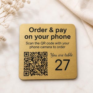 Op de afbeelding: Een vierkant goudkleurig metalen bordje met afgeronde hoeken en zwarte tekst die luidt Order & pay on your phone, Scan the QR code with your phone camera to order, You are table, het getal 27 en een zwarte QR-code op de metalen ondergrond.