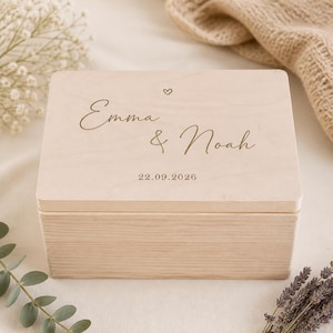Op de afbeelding: Een rechthoekige bewaardoos van lichtgekleurd hout met een gladde afwerking. Het deksel is gegraveerd met de namen Emma & Noah in een cursief lettertype, een klein hartsymbool boven de namen en de datum 22.09.2026 onder de namen.