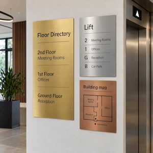 Op de afbeelding: Een set van drie rechthoekige metalen borden gemonteerd op een muur. Een hoog goudkleurig bord vermeldt Floor Directory, 2nd Floor Meeting Rooms, 1st Floor Offices en Ground Floor Reception. Ook een zilveren Lift-bord en een koperen Building map.