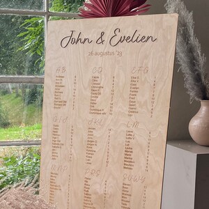 Può includere: Un tableau di posti a sedere in legno con i nomi degli ospiti elencati in ordine alfabetico. Il tableau è decorato con un ventaglio color borgogna e erba della pampa beige. Il testo in alto recita "John & Evelien 26 agosto '23" e in basso "Trova il tuo tavolo".