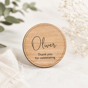 Op de afbeelding: Een ronde houten schijf met een natuurlijke lichtbruine houtnerf. De tekst "Oliver" is gegraveerd in een zwart scriptlettertype, gevolgd door de tekst "Thank you for celebrating" in een eenvoudig, strak zwart schreefloos lettertype.