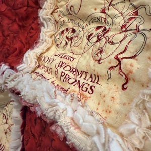 May include: Close-up of a fabric patchwork with cream, red, and white colors. The cream-colored fabric has red text and illustrations. The red fabric has a textured appearance. White ruffled fabric borders the patchwork.