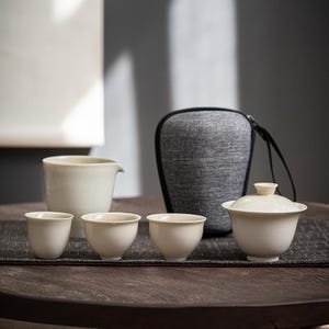 Pode incluir: Um conjunto de chá composto por três pequenas chávenas de cerâmica em tom creme, um jarro a condizer com bico e uma taça com tampa. O conjunto é acompanhado por um estojo de transporte cinzento e texturado com fecho e pega preta.