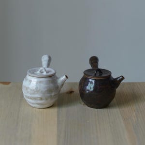 Peut inclure: Deux petites théières en céramique avec poignées latérales et couvercles. L'une est dotée d'un vernis blanc brillant avec des stries horizontales, tandis que l'autre présente un vernis brun foncé moucheté et une forme arrondie texturée.