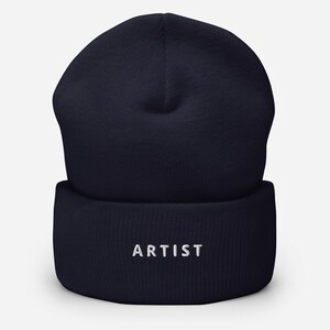 May include: A navy blue beanie hat with the word "ARTIST" embroidered in white. The hat is made of a soft, knitted material and has a folded brim. The beanie is a classic style, suitable for cold weather.
