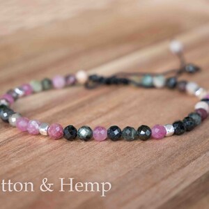 May include: A delicate bracelet made with pink, green, and black gemstone beads and silver accents. The bracelet is tied with a hemp cord.