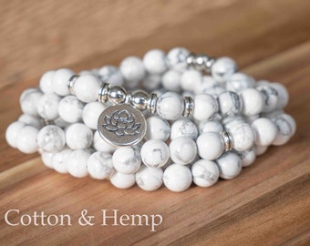 May include: White Howlite gemstone mala beads with a silver lotus charm. The mala is made of multiple strands of beads and is wrapped around a wooden surface.