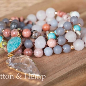 May include: A multi-strand beaded necklace with a clear quartz point pendant and a teal green gemstone. The necklace features various shades of gray, pink, and brown beads.