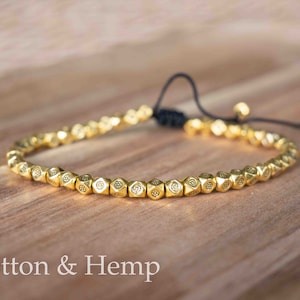 May include: A gold beaded bracelet with a black cord closure. The beads are small and have a geometric design.