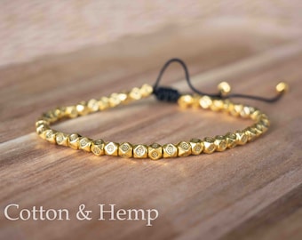 May include: A gold beaded bracelet with a black cord closure. The beads are small and have a geometric design.