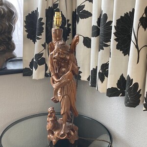 May include: A wooden lamp with a carved figure of an elderly man holding a fish, and a small monkey at the base. The lamp has a gold bulb socket and a brown cord. The lamp is on a glass table.