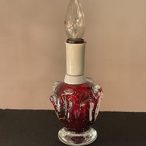 Puede incluir: Una lámpara decorativa de cristal rojo con una bombilla transparente. La lámpara tiene una base bulbosa con detalles de cristal transparente y un cuello blanco. La bombilla es transparente y puntiaguda.