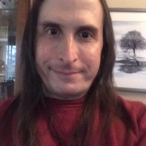 Puede incluir: Un hombre con cabello largo y castaño lleva una sudadera roja con un collar negro y un colgante redondo. El colgante tiene un diseño blanco.