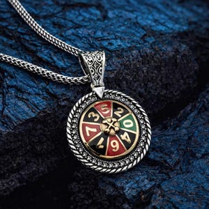 May include: Silver-toned pendant necklace featuring a roulette wheel design. The pendant has a black, red, and green color scheme with numbers. The necklace is displayed on a textured, dark blue surface.