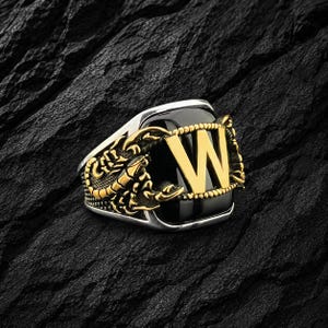 May include: Silver ring with a black onyx stone, featuring a gold scorpion design and a gold letter "W". The ring is set against a dark, textured background.