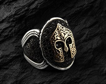 May include: A silver ring with a black stone setting and a gold-colored helmet design. The ring has intricate detailing on the band and the helmet, showcasing a blend of textures and colors. The ring is a statement piece.