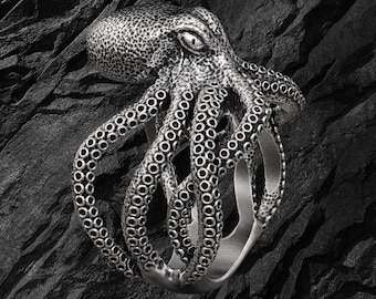 May include: A silver-toned ring featuring a detailed octopus design. The octopus head is textured with small bumps, and its tentacles wrap around the band, displaying visible suction cups. The metal has a dark, oxidized finish in the crevices.