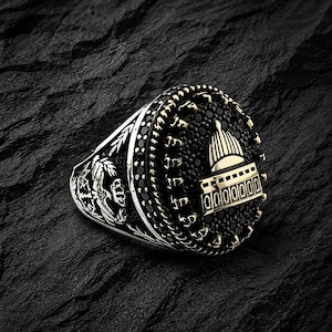May include: A silver ring with a black stone setting and gold accents. The ring features a detailed design of a building with a dome, surrounded by a decorative border. The ring is set against a dark, textured background.