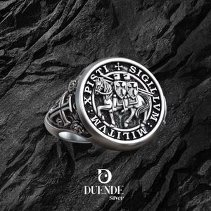 Könnte beinhalten: Ein silberner Ring mit detailliertem Design, das einen Ritter zu Pferd, ein Kreuz und den Text "XPISTI SIGILLVM MILITVM" zeigt. Der Ring hat eine runde Oberfläche und aufwendige Seitendetails. Der Markenname "DUENDE Silver" ist ebenfalls sichtbar.