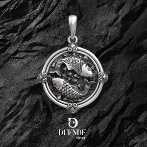 Puede incluir: Colgante de plata con dos peces dentro de un marco circular con detalles ornamentales. El colgante tiene un asa para una cadena y est&aacute; sobre un fondo oscuro y texturizado. La marca "DUENDE Silver" se muestra en la parte inferior.