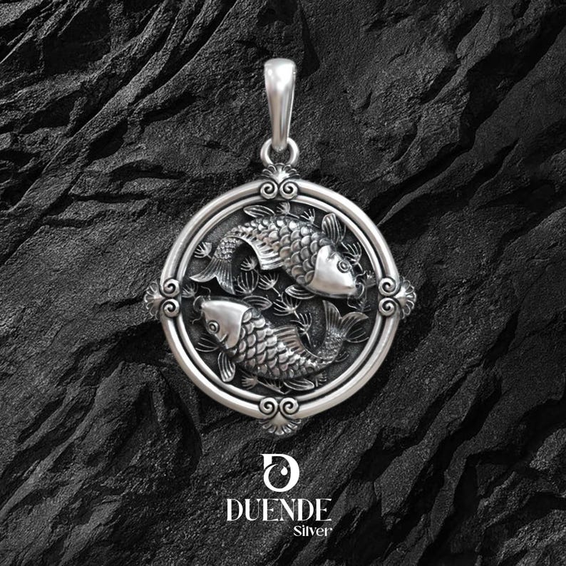 Puede incluir: Colgante de plata con dos peces dentro de un marco circular con detalles ornamentales. El colgante tiene un asa para una cadena y est&aacute; sobre un fondo oscuro y texturizado. La marca "DUENDE Silver" se muestra en la parte inferior.