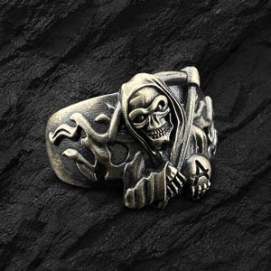 May include: A detailed, antique-style ring featuring a grim reaper design. The ring is cast in a bronze-toned metal and depicts the grim reaper holding a scythe, with a skull face and hooded cloak. The ring has a detailed, textured finish.