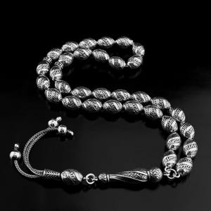 Può includere: Una collana di perline di preghiera color argento con un design intricato e strutturato. Le perline ovali sono infilate insieme, con un nappa e un ciondolo decorativi. Lo sfondo è nero.