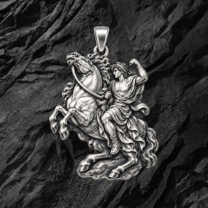 May include: A silver-toned pendant featuring a detailed relief of a figure in flowing robes riding a rearing horse. The design includes a prominent bail at the top for attachment. The metal surface has an oxidized finish to highlight the intricate textures of the horse's mane, the rider's musculature, and the fabric folds.