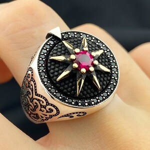 May include: A silver ring with a starburst design. The ring features a central red gemstone surrounded by gold-colored points and a border of black stones. Intricate scrollwork adorns the side of the ring, creating a detailed and elegant appearance.