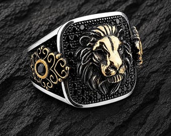 May include: A silver ring with a gold lion's head centerpiece. The lion's mane and face are gold, set against a black background of small stones. The ring has ornate gold detailing on the sides and a black stone accent.